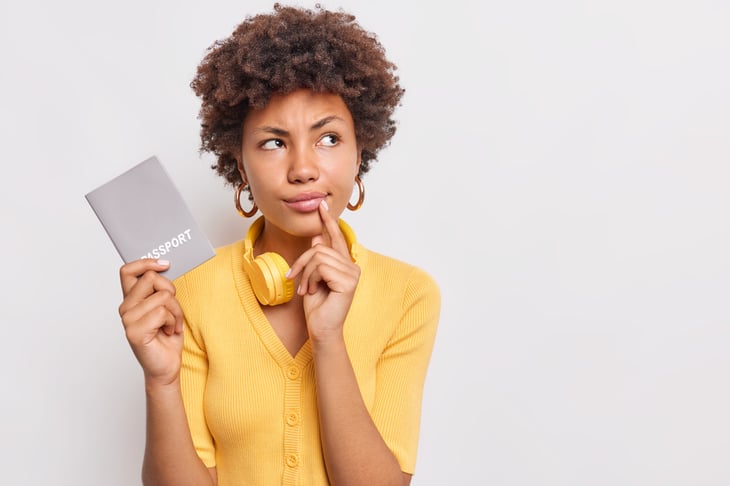 serious-thoughtful-woman-with-afro-hair-considers-future-travel-looks-pensively-away-wears-casual-yellow-jumper-holds-passport-official-document-poses-against-white-wall